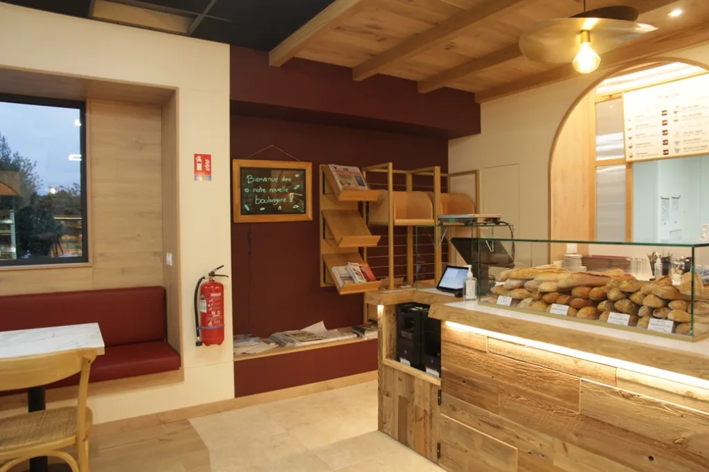 Coin salon et caisse d'Augustin Guingamp : banquette bordeaux, présentoir de presse et comptoir en bois vieilli garni de pains