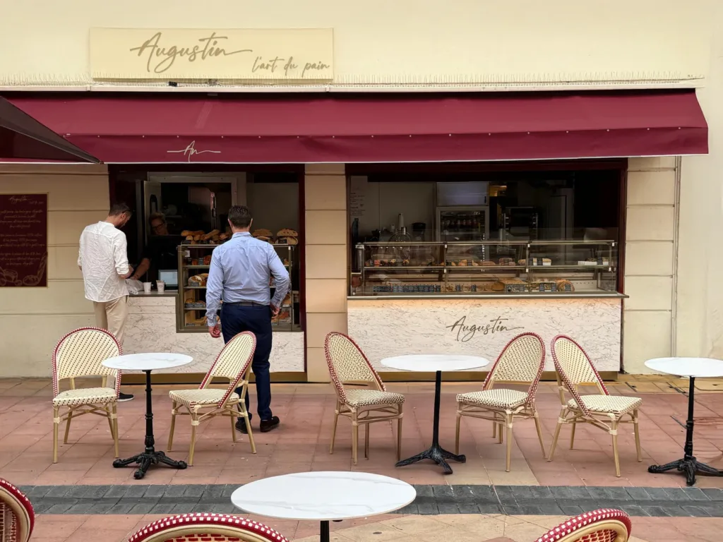 Clients au comptoir d'Augustin Menton devant les pains et les viennoiseries, sous l'enseigne « Augustin – L'art du pain »